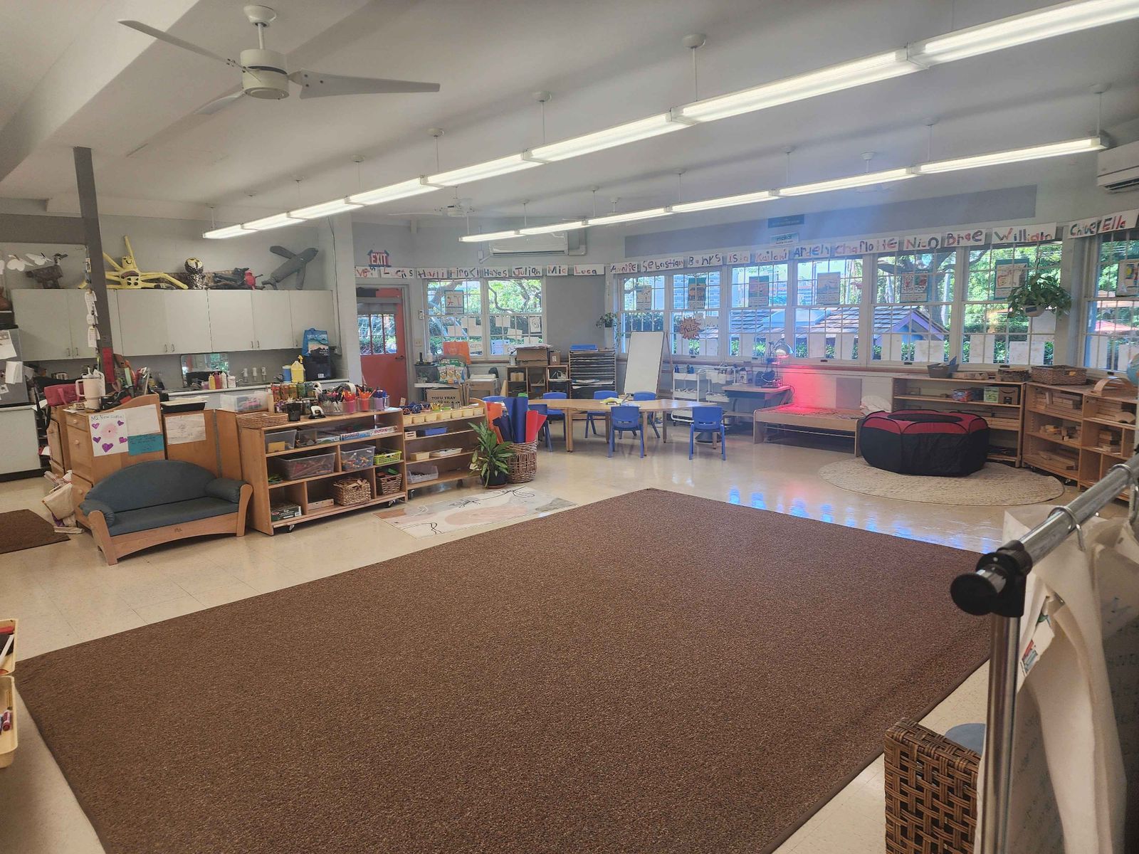 A wide classroom with a large brown rug, low wooden shelves, small tables and chairs, a child-sized couch, and a row of children's names above the windows.