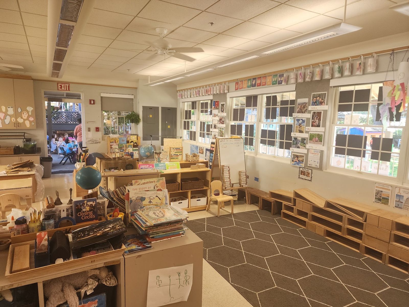 A bright early-grades classroom with wooden shelves of books and supplies, low wooden benches, and family photos taped in the windows.