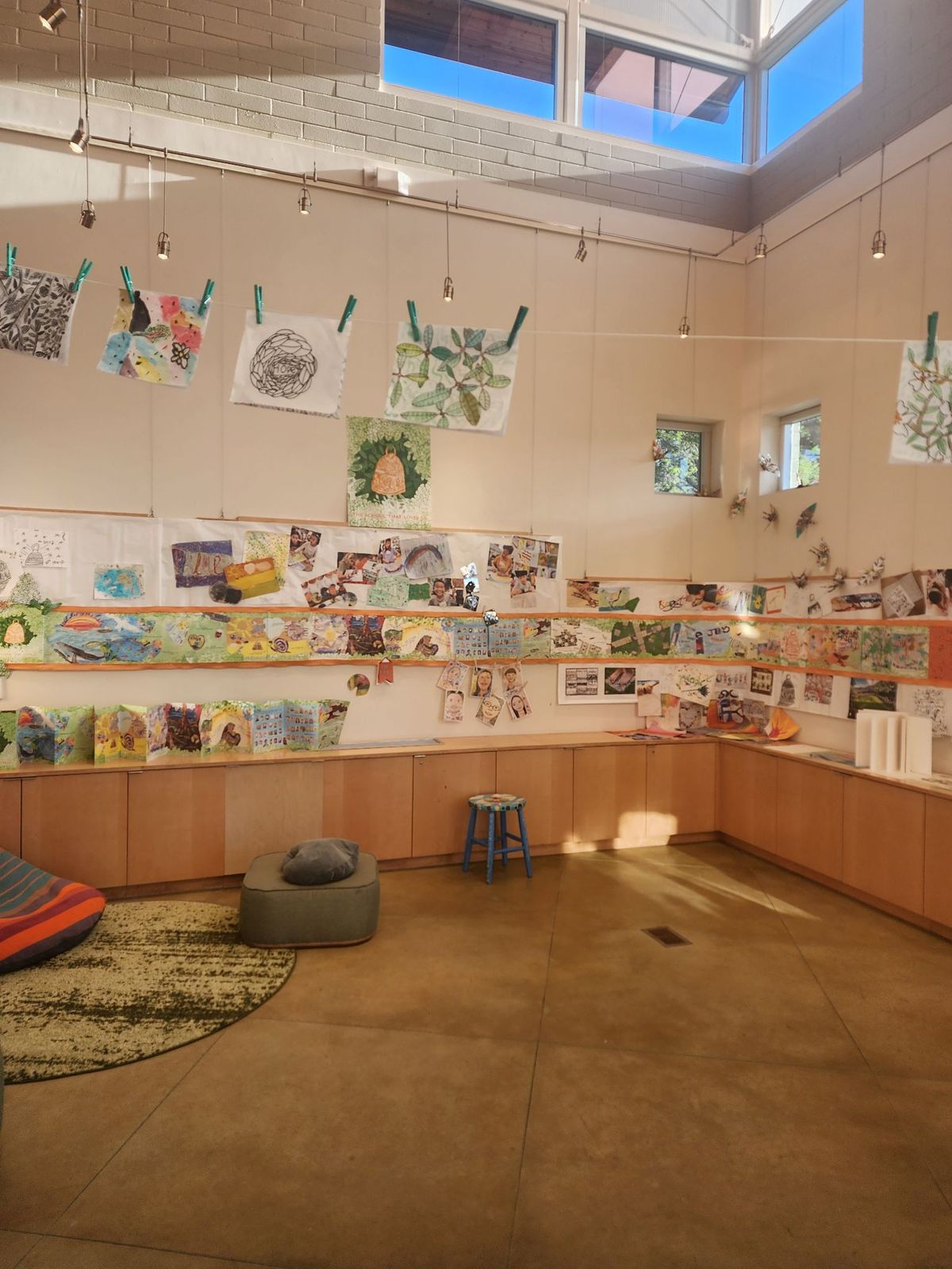 A high-ceilinged gallery room with student paintings and prints clipped to wires and lining long shelves, with a rug and floor cushions below.