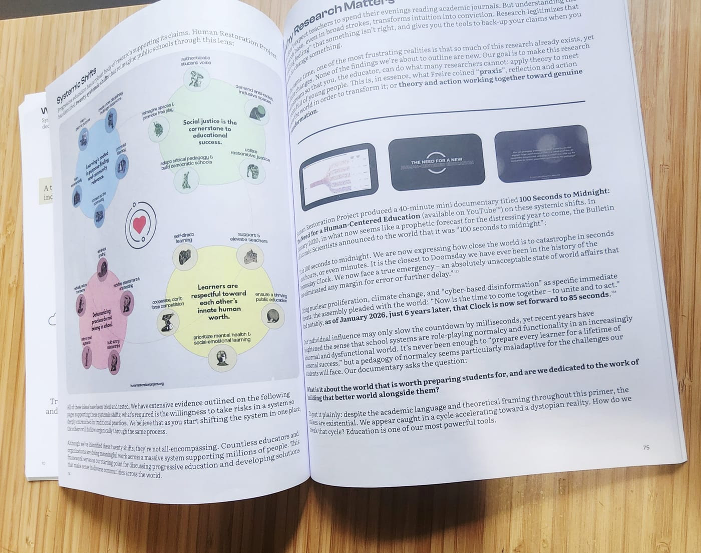 A printed copy of We Are Worldbuilders open on a wooden table, showing an illustrated "Systemic Shifts" diagram on the left page and a "Why Research Matters" essay on the right.